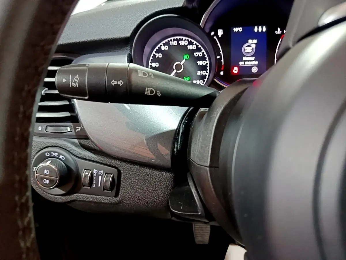 Gros plan sur le combiné d'instruments et commandes de phares à gauche du volant d'une FIAT 500X gris foncé, 2019.