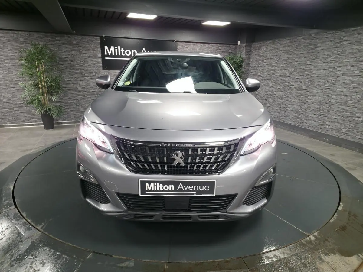 Vue avant frontale d'un Peugeot 3008 gris métallisé avec calandre noire et phares allumés en intérieur showroom.