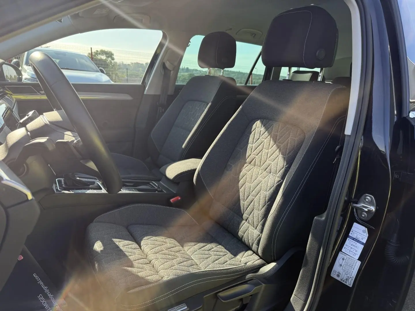Vue intérieure côté conducteur de la Volkswagen Passat SW 2023, sièges tissu noir avec surpiqûres blanches et accoudoir central.