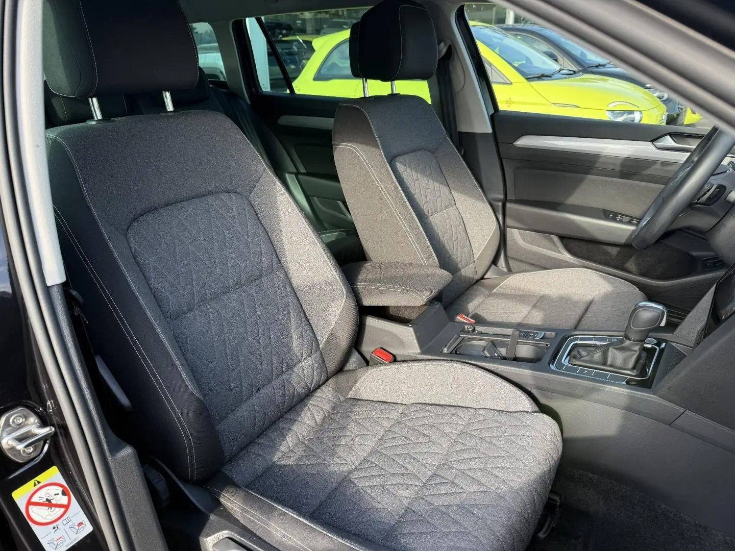 Vue intérieure côté conducteur de la Volkswagen Passat SW 2023, sièges tissu gris avec accoudoir central noir.