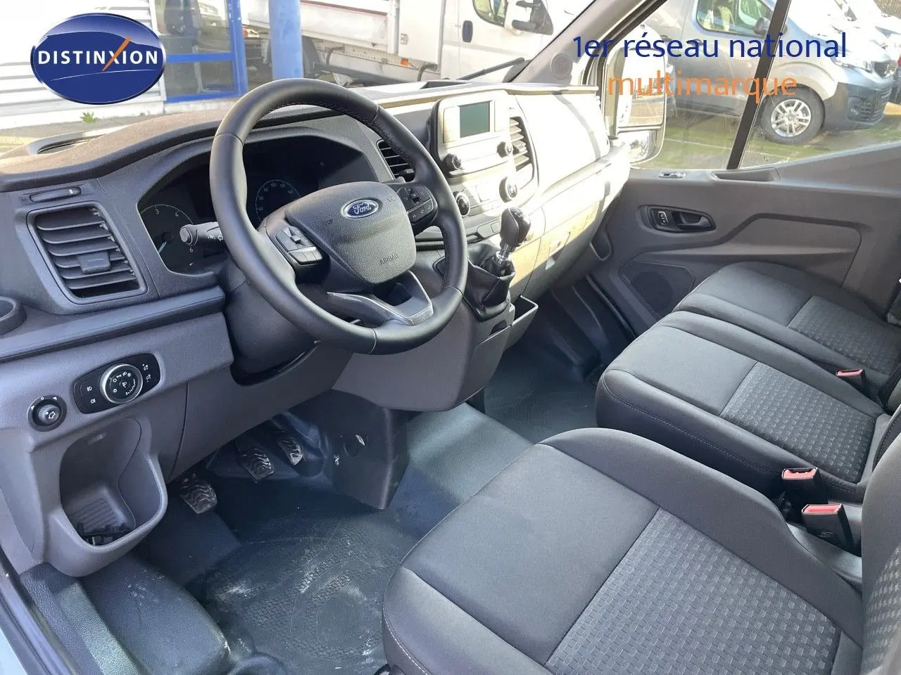 Vue intérieure côté conducteur du Ford Transit Fourgon blanc 2023, montrant le tableau de bord et les sièges en tissu noir.