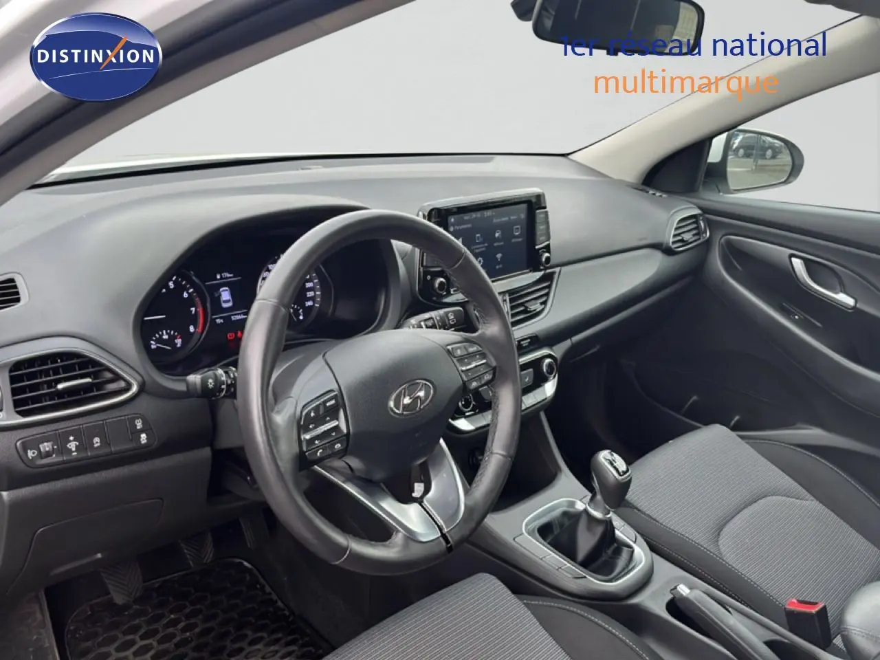 Vue intérieure côté conducteur de la Hyundai i30 2023, tableau de bord moderne avec écran tactile et levier de vitesses manuel.