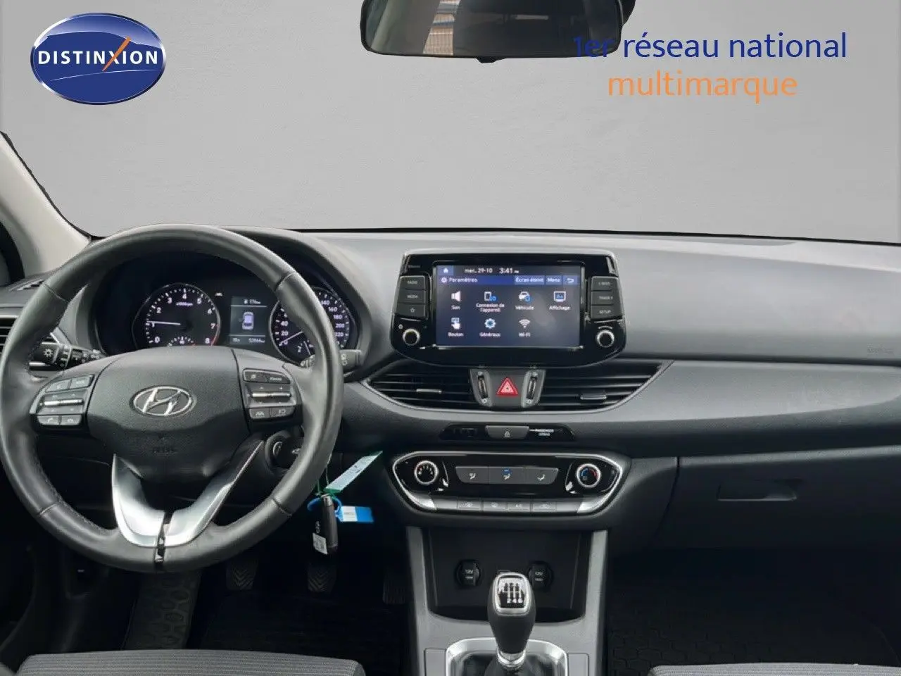 Vue intérieure frontale du tableau de bord et volant de la Hyundai i30 blanche nacrée, avec écran tactile et boîte manuelle.