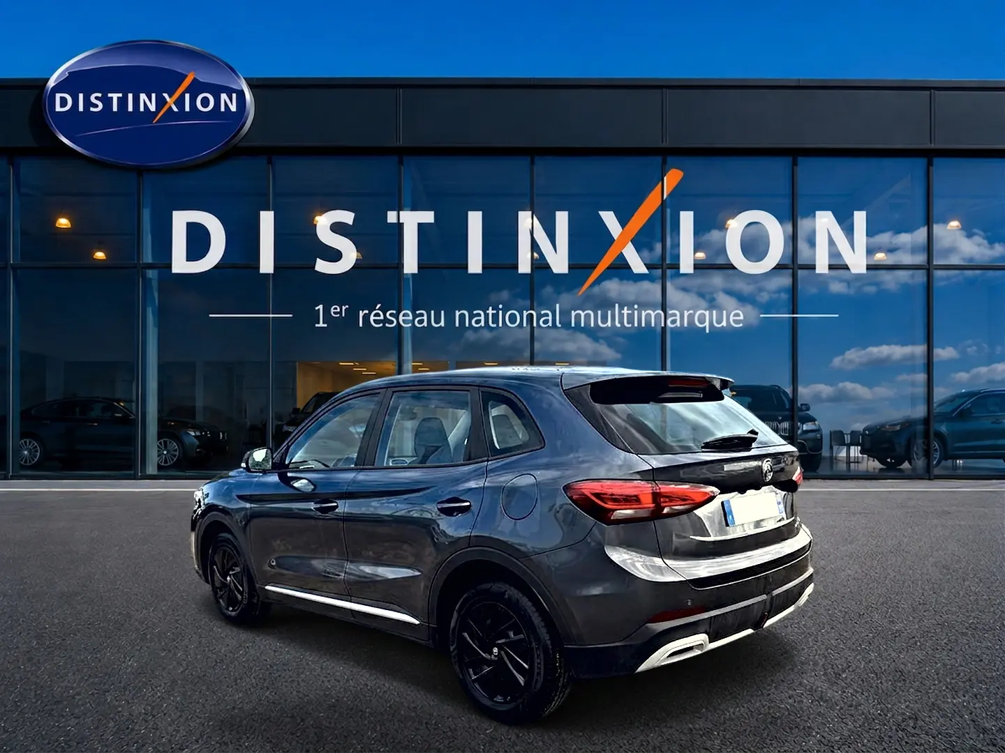 Vue 3/4 arrière droite du MG ZS Camden grey 2026 avec feux LED et jantes noires devant un showroom Distinxion.
