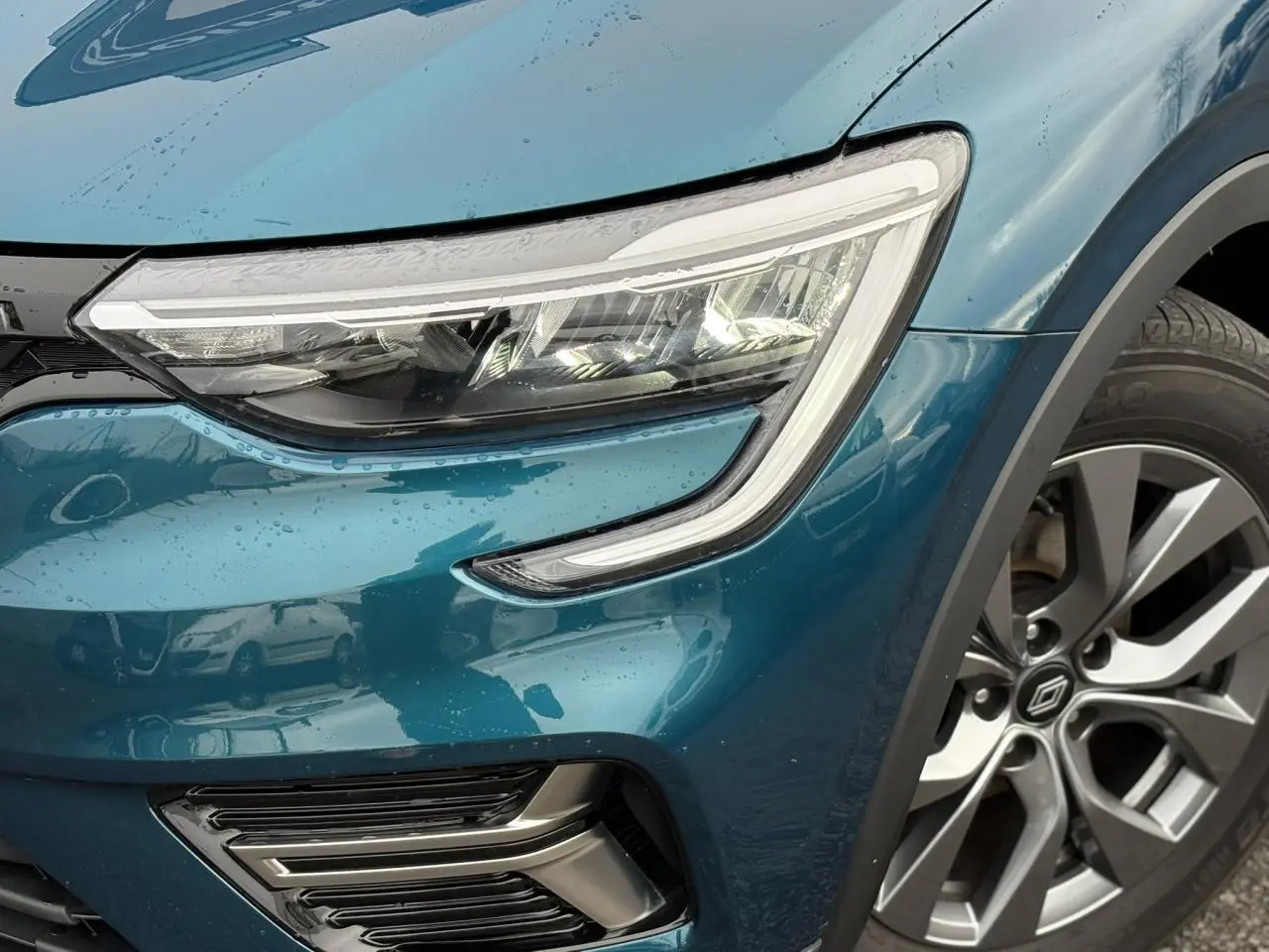 Gros plan sur le phare avant droit et la roue alliage grise du Renault Arkana Bleu Zanzibar sous la pluie.