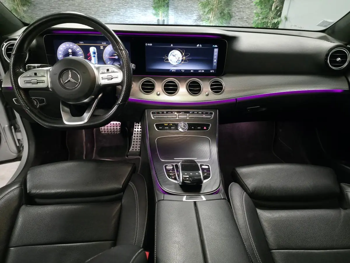 Vue intérieure avant de la Mercedes Classe E Break 2020, tableau de bord noir avec éclairage d'ambiance violet et volant multifonction.