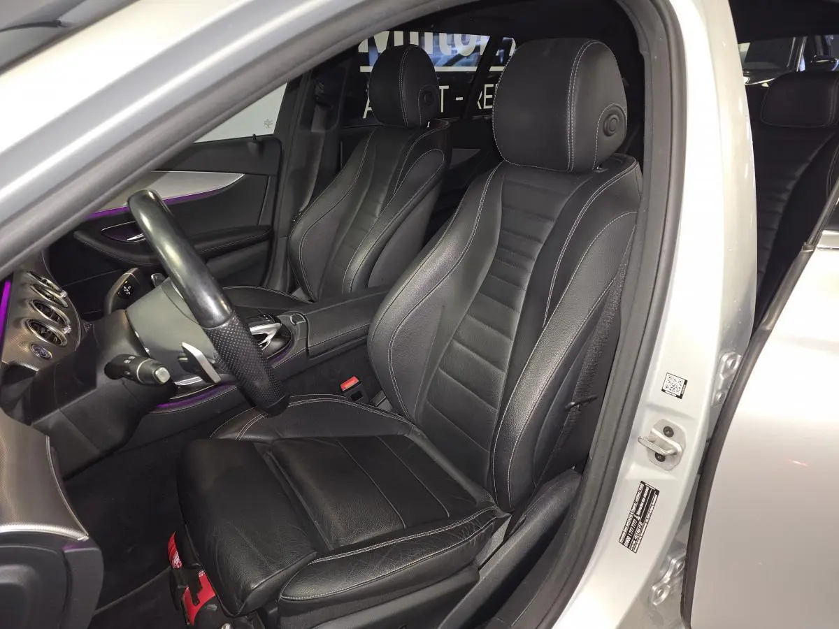 Vue intérieure côté conducteur d'une Mercedes Classe E Break gris, sièges cuir noir avec surpiqûres blanches et éclairage d'ambiance violet.
