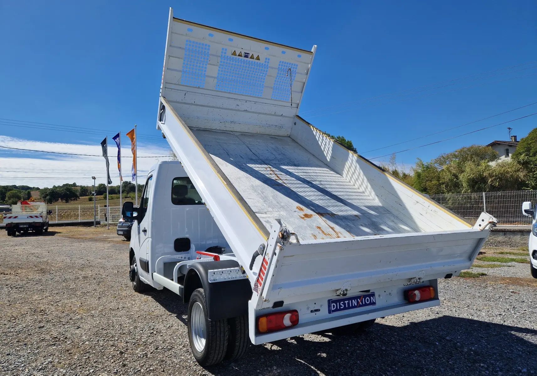 Renault Master Benne blanc vu en 3/4 arrière avec benne relevée et roues jumelées sur terrain extérieur.