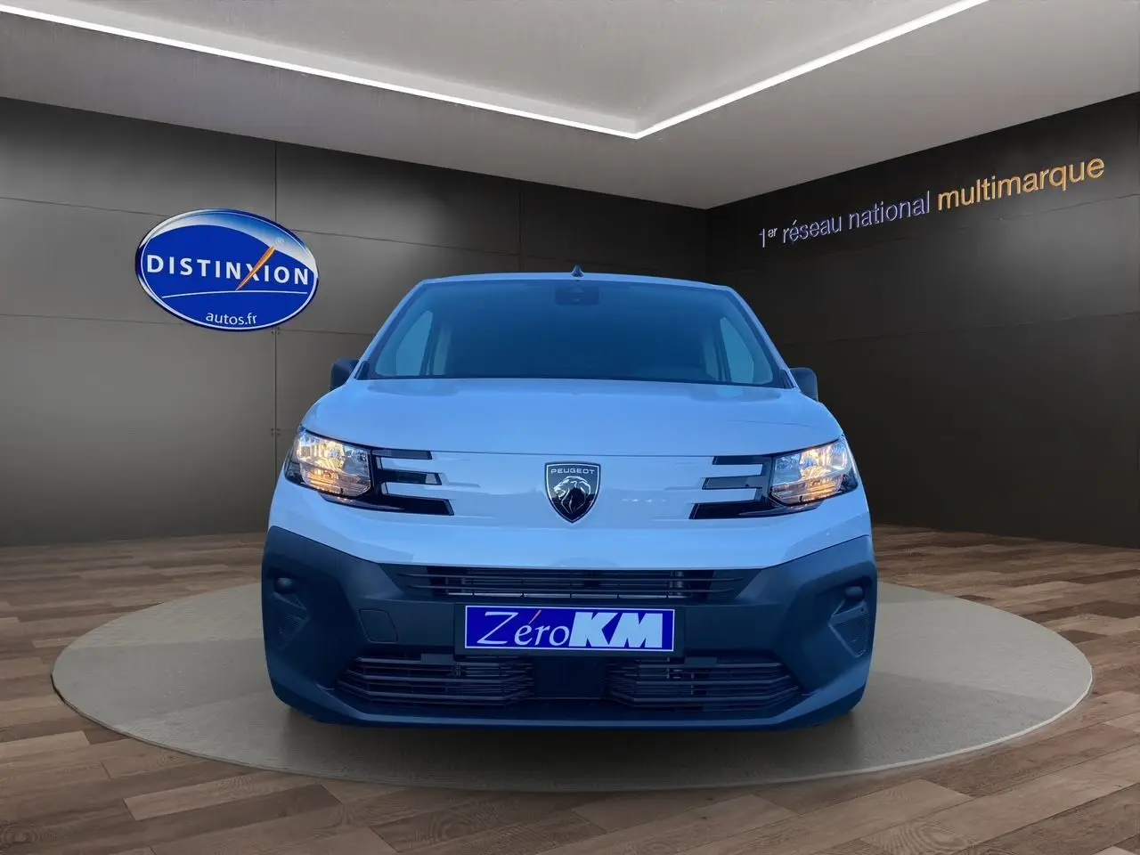 Vue frontale d'un Peugeot Partner blanc 2024 avec phares allumés et plaque "Zéro KM" en concession.