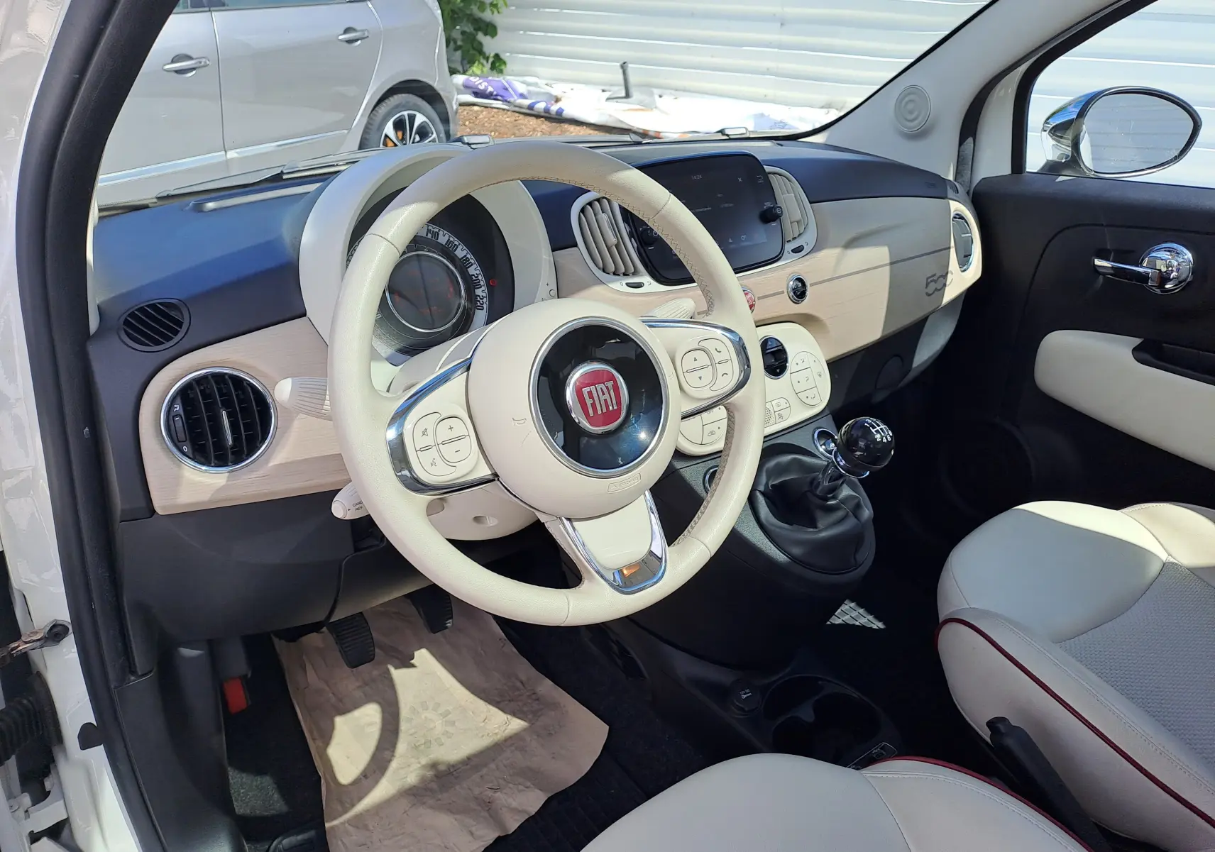 Intérieur de Fiat 500 blanc, vue côté conducteur, volant beige, tableau de bord clair avec écran tactile et levier de vitesses manuel.