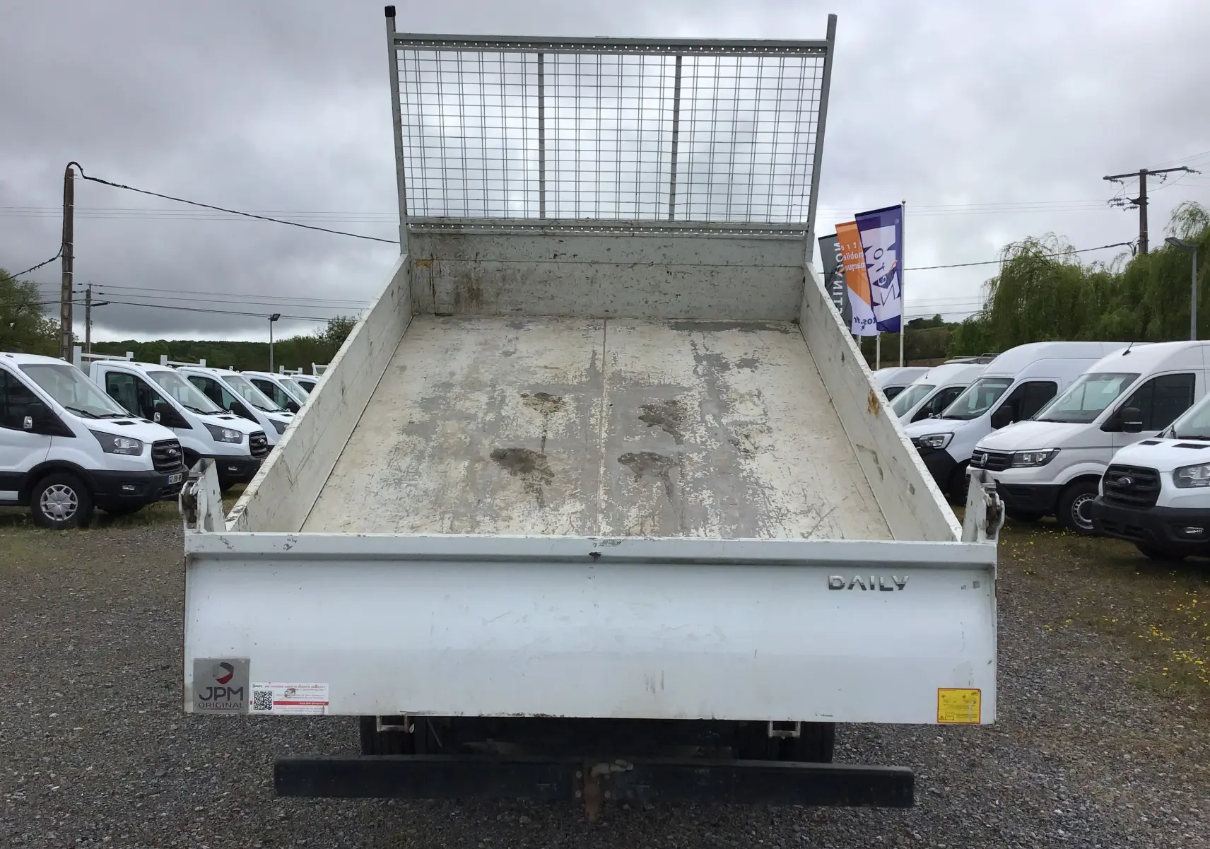 Vue arrière d'un Iveco Daily blanc avec benne basculante et grille de protection métallique.