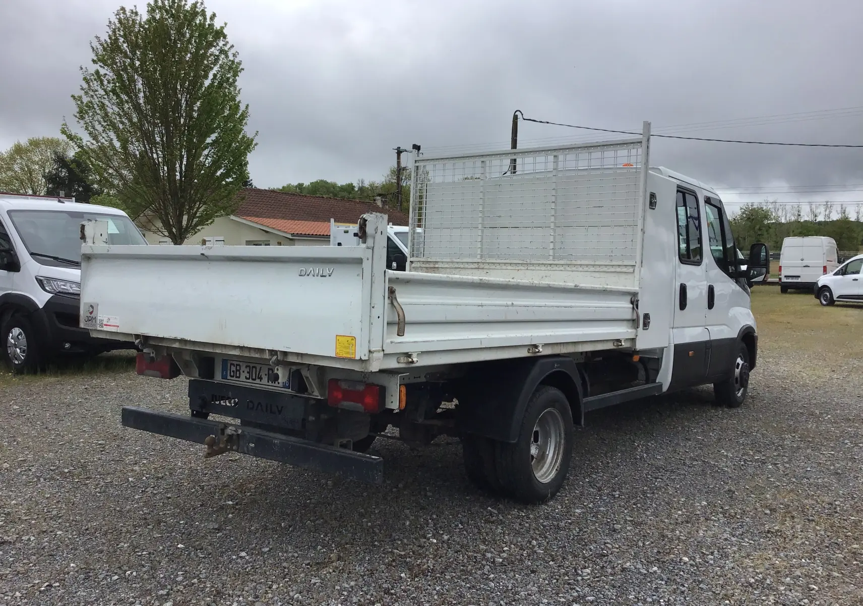 IVECO Daily blanc vue 3/4 arrière droit, benne avec grille et roues jumelées sur sol gravillonné.