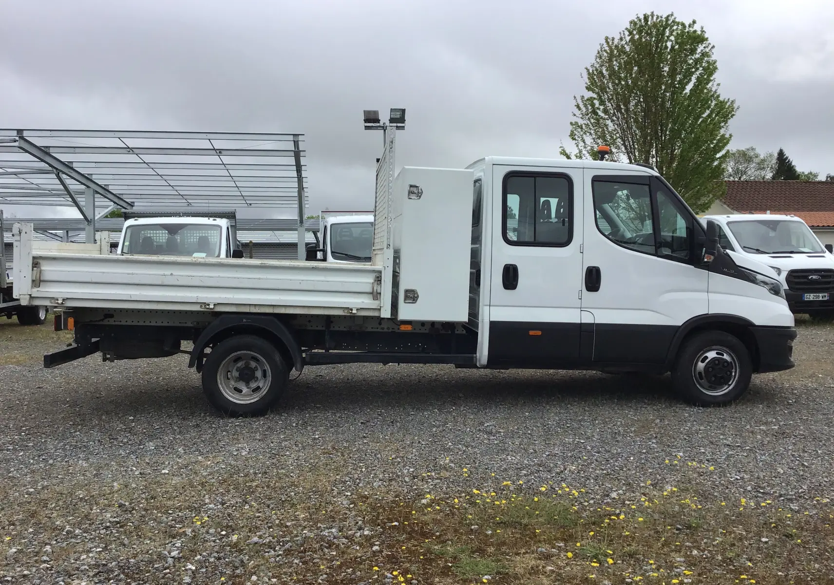 Vue de profil côté gauche d'un Iveco Daily blanc avec benne, coffre latéral et cabine double sur un sol gravillonné.