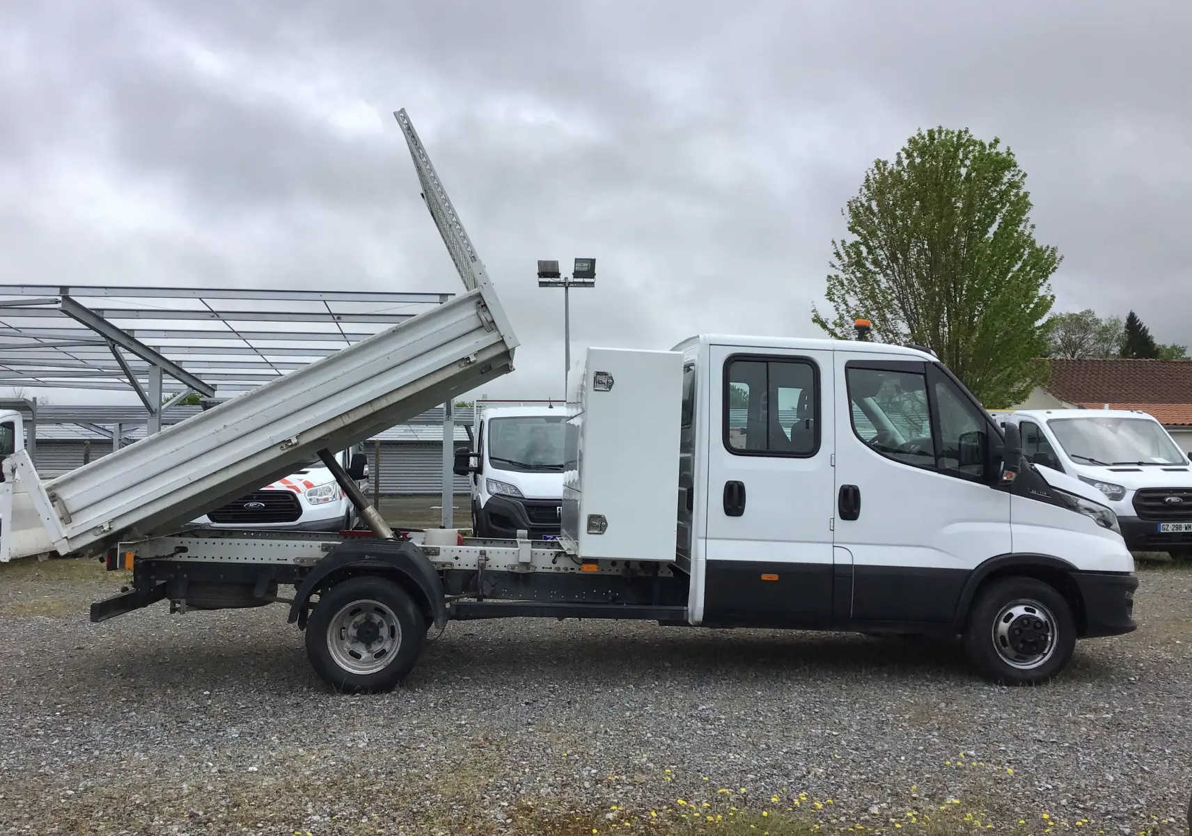 Profil droit d'un Iveco Daily blanc avec benne basculante relevée et coffre latéral sur un terrain extérieur.