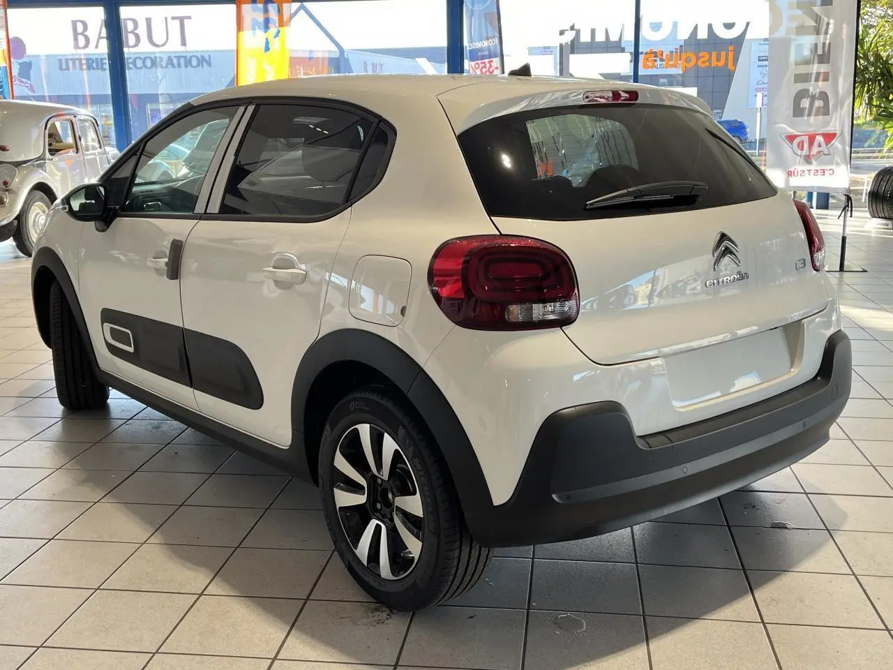 Citroën C3 blanc vue 3/4 arrière droit en showroom, avec protections noires et jantes alliage bicolores.