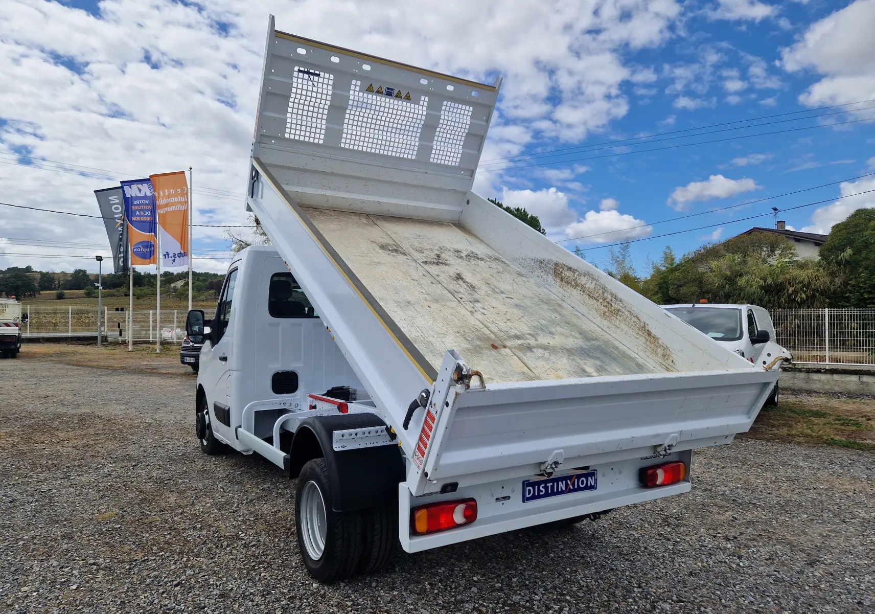 Vue 3/4 arrière droite d’un Renault Master blanc avec benne relevée et roues jumelées sur un sol gravillonné.