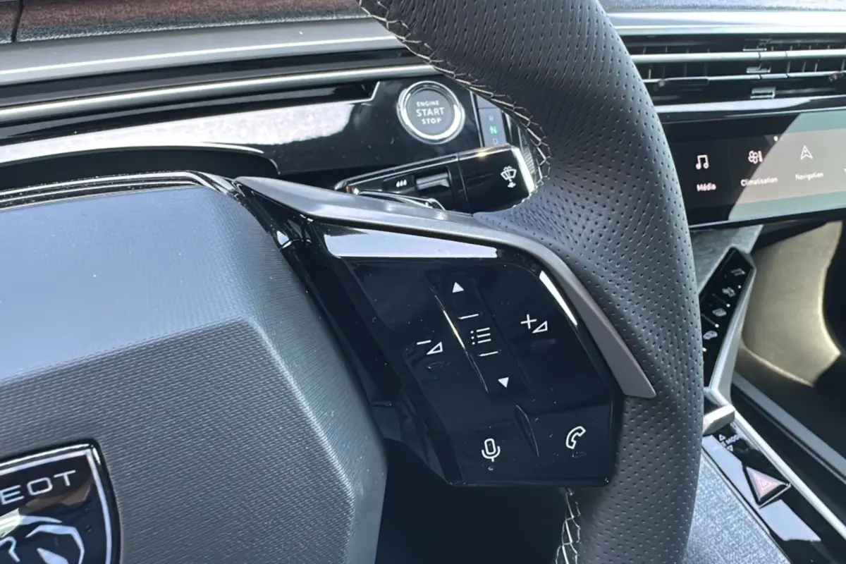 Gros plan sur le volant perforé du Peugeot 3008 Hybrid 2025 avec commandes intégrées et bouton démarrage visible.
