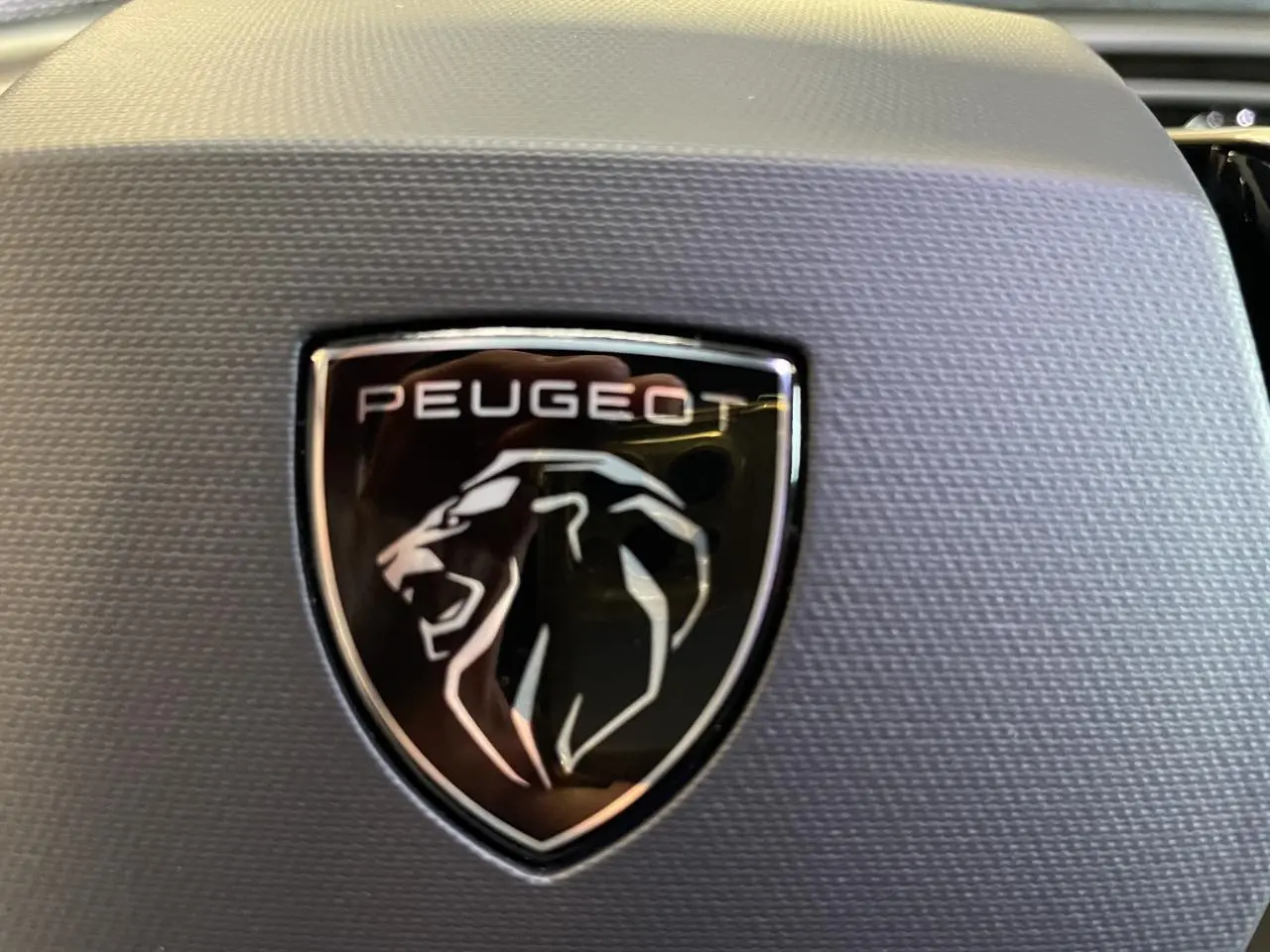 Gros plan sur le logo Peugeot noir et argenté sur fond texturé gris, intérieur du volant du 3008 GT hybride blanc.