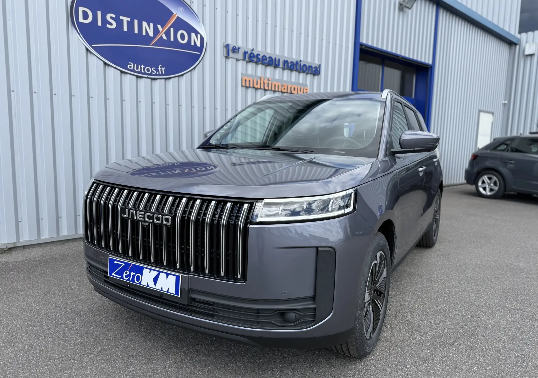 Vue 3/4 avant du JAECOO J5 gris Phantom avec calandre verticale noire et phares LED fins, stationné devant un bâtiment.