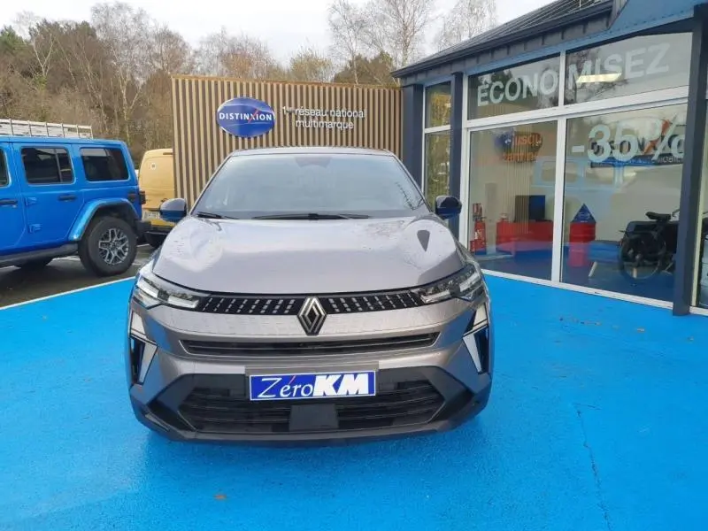 Vue avant d'un Renault Captur 2025 gris Cassiopée avec calandre noire et logo Renault centré, stationné sur sol bleu.