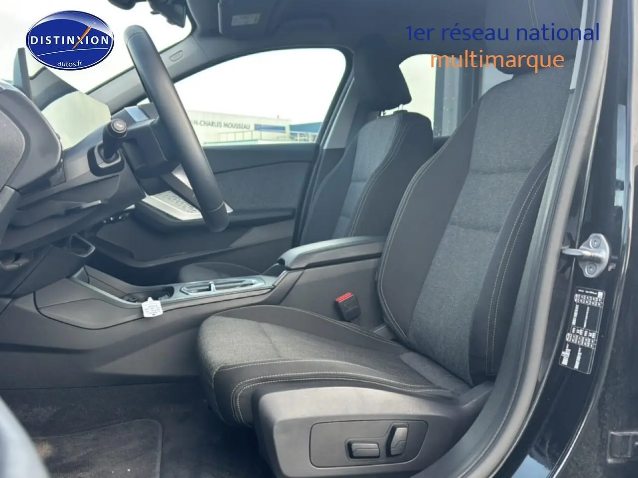 Vue intérieure côté conducteur de la BMW Série 1 120da 2024 noire, sièges tissu anthracite avec accoudoir central.