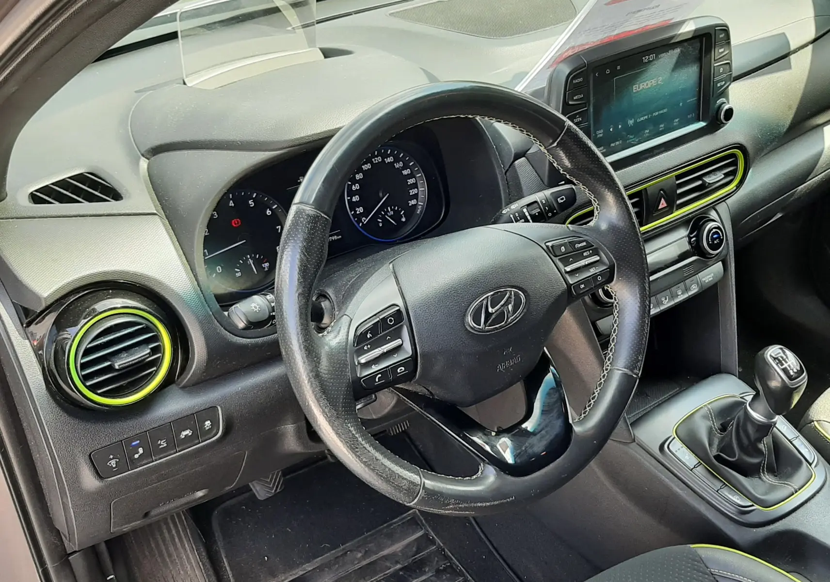 Vue intérieure côté conducteur du Hyundai Kona gris 2018, volant cuir, tableau de bord avec écran tactile et levier de vitesse manuel.