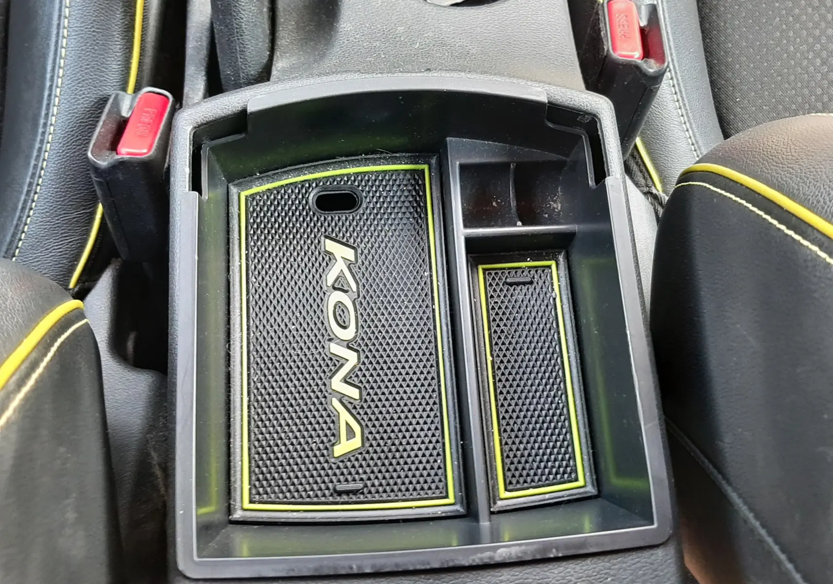 Vue plongeante sur le rangement central intérieur du Hyundai Kona gris avec tapis antidérapant marqué "KONA" et surpiqûres jaunes.
