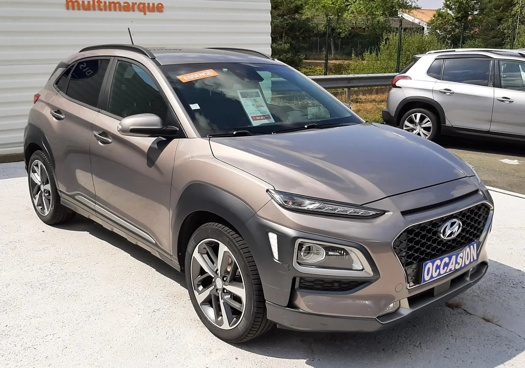 Hyundai Kona gris en 3/4 avant droit, avec calandre noire et jantes alliage bicolores visibles.