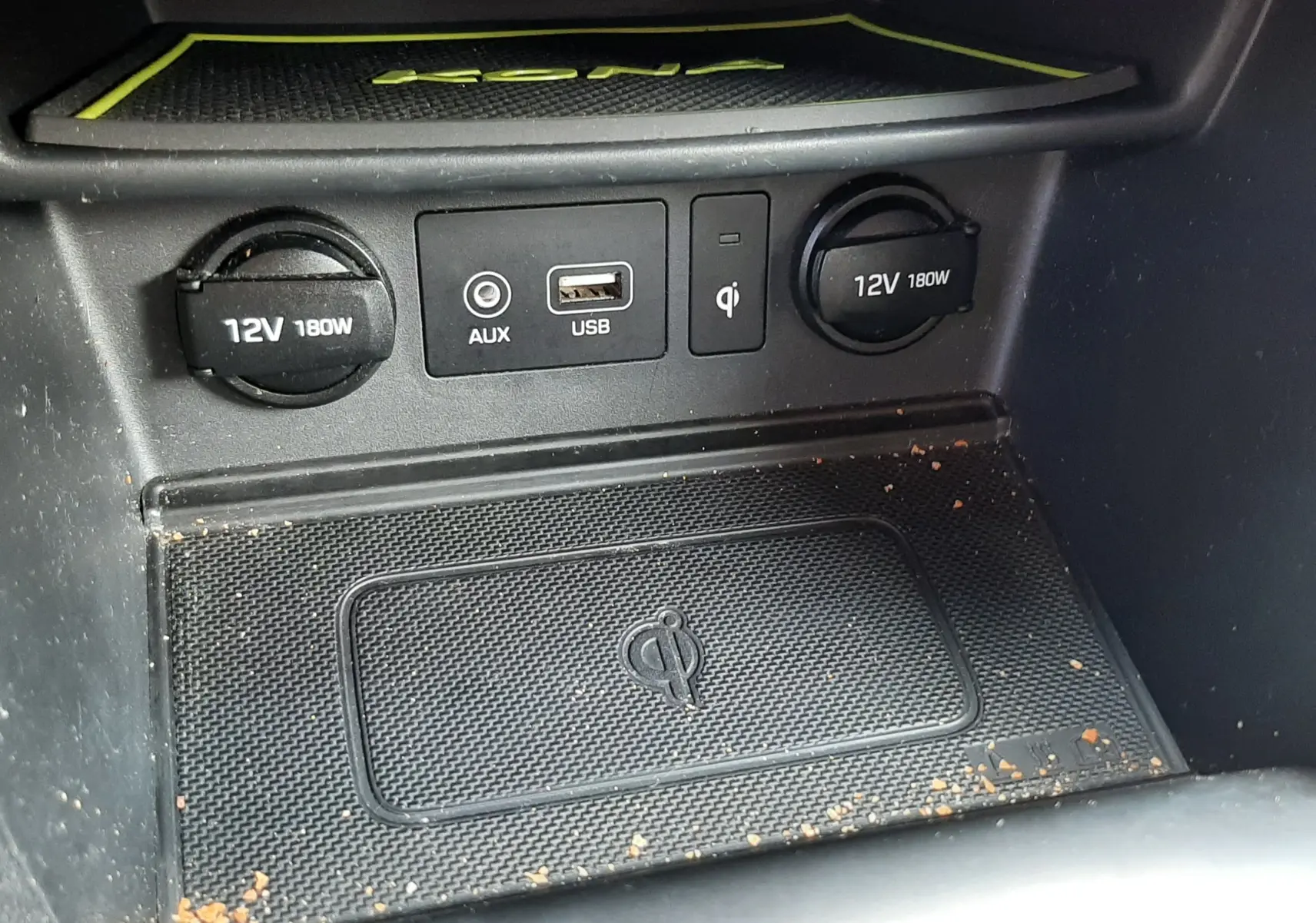 Gros plan sur la console centrale du Hyundai Kona gris, montrant prises 12V, USB, AUX et chargeur sans fil Qi avec traces de saleté.