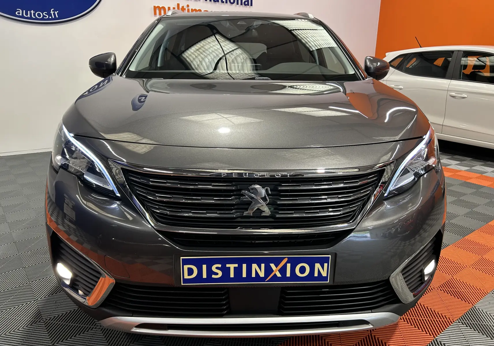 Vue de face d'un Peugeot 5008 gris métallisé avec calandre chromée et feux LED allumés en intérieur showroom.