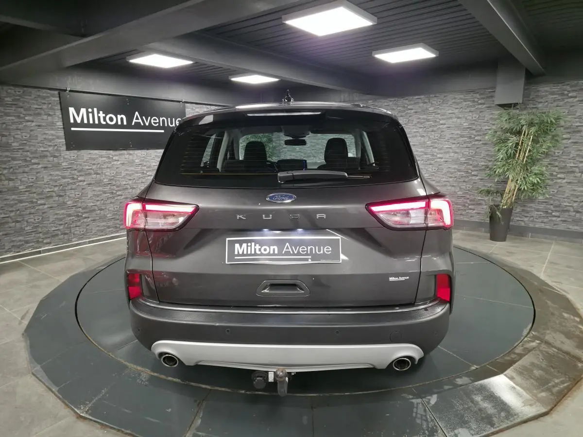 Vue arrière d'un Ford Kuga gris métallisé 2020 avec attelage fixe et feux arrière LED allumés dans un showroom.