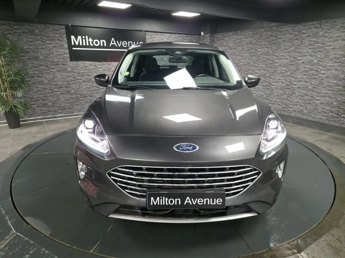 Vue avant d'un Ford Kuga gris métallisé 2020 avec calandre chromée et phares allumés dans un showroom.
