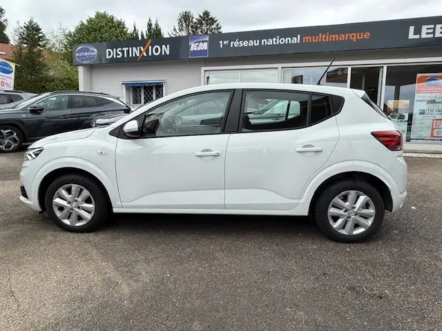 Profil côté gauche d'une Dacia Sandero blanc glacier 2023 garée devant un concessionnaire multimarque.