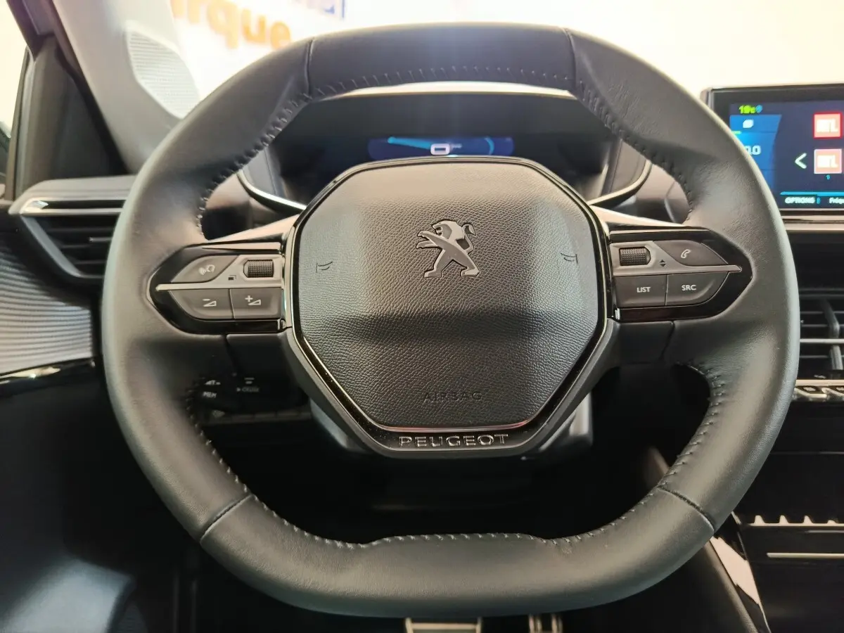Gros plan sur le volant cuir noir multifonctions de la Peugeot 208 électrique avec tableau de bord numérique en arrière-plan.