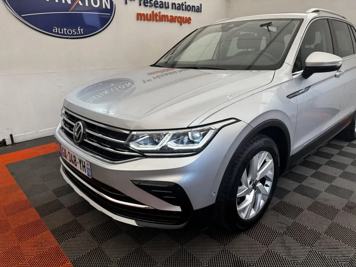 Vue 3/4 avant droit d’un Volkswagen Tiguan gris clair avec calandre chromée et phares LED allumés en intérieur showroom.