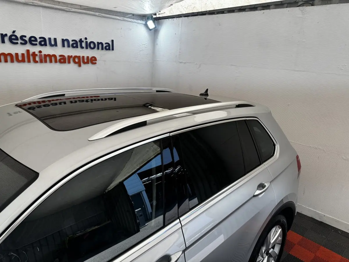 Vue 3/4 arrière droite d’un Volkswagen Tiguan gris clair avec toit ouvrant et barres de toit chromées.