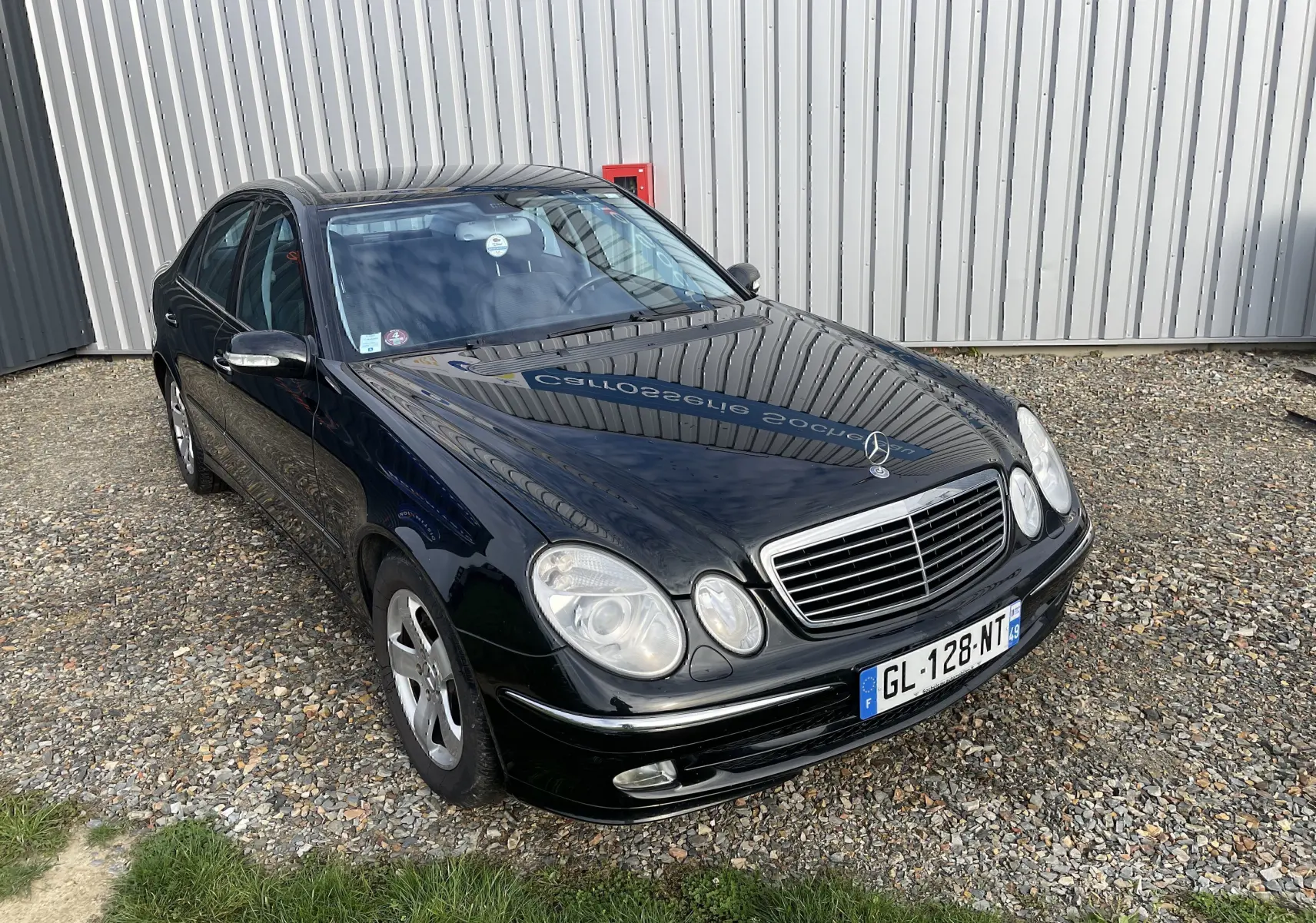 Mercedes Classe E 320 CDI Elegance noire vue en 3/4 avant sur un sol gravillonné devant un bâtiment métallique.