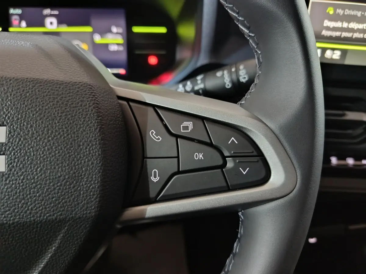 Gros plan sur les commandes multifonctions à droite du volant gris foncé du Dacia Bigster, avec affichage numérique flou en arrière-plan.