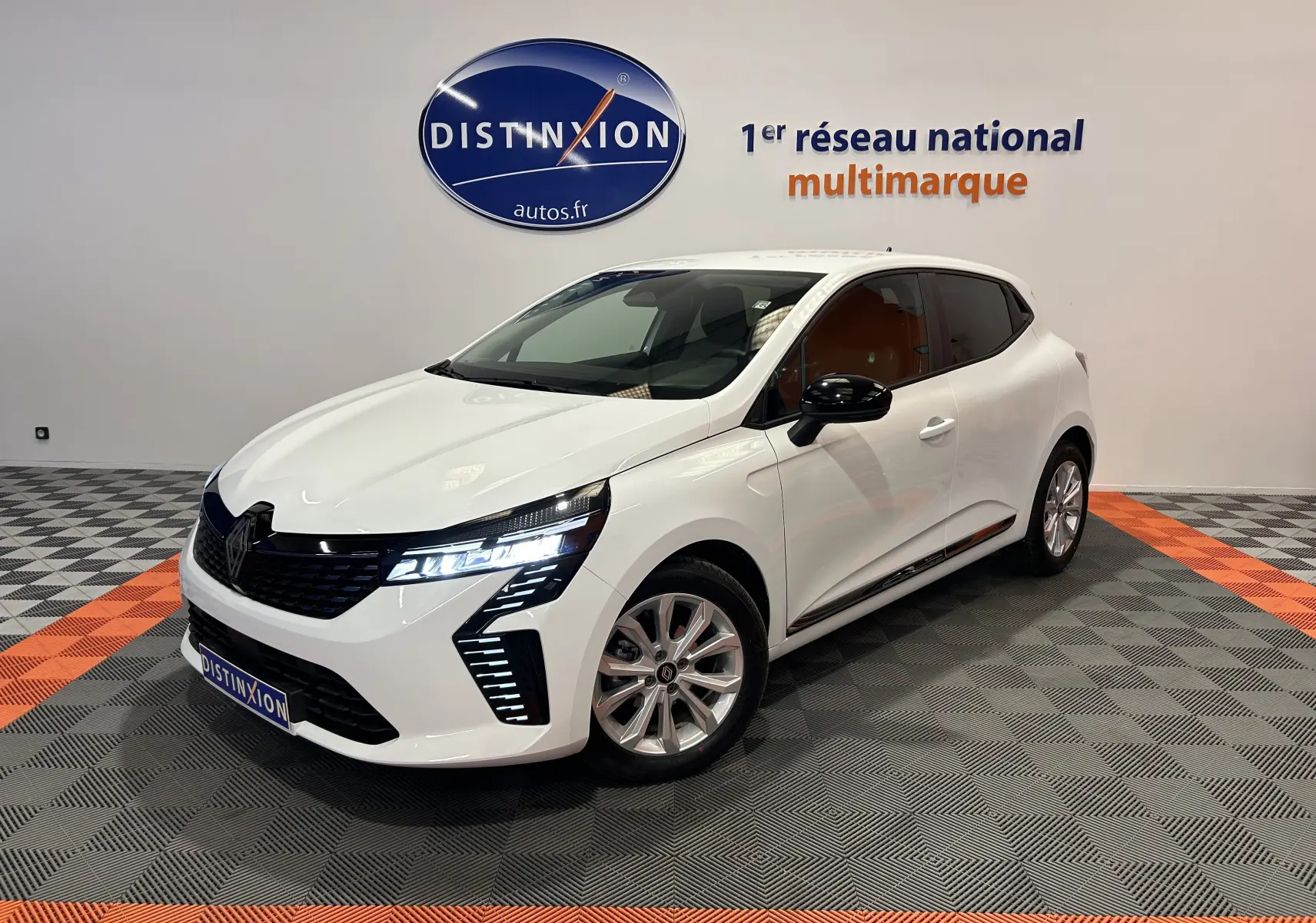 Renault Clio V blanche vue 3/4 avant droit avec feux LED allumés dans un showroom multimarque.
