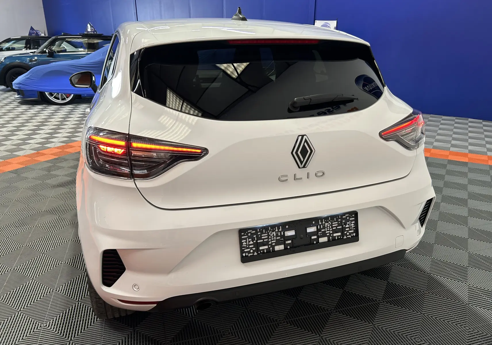 Vue arrière d'une Renault Clio V blanche avec feux allumés, stationnée dans un showroom au sol à damier.