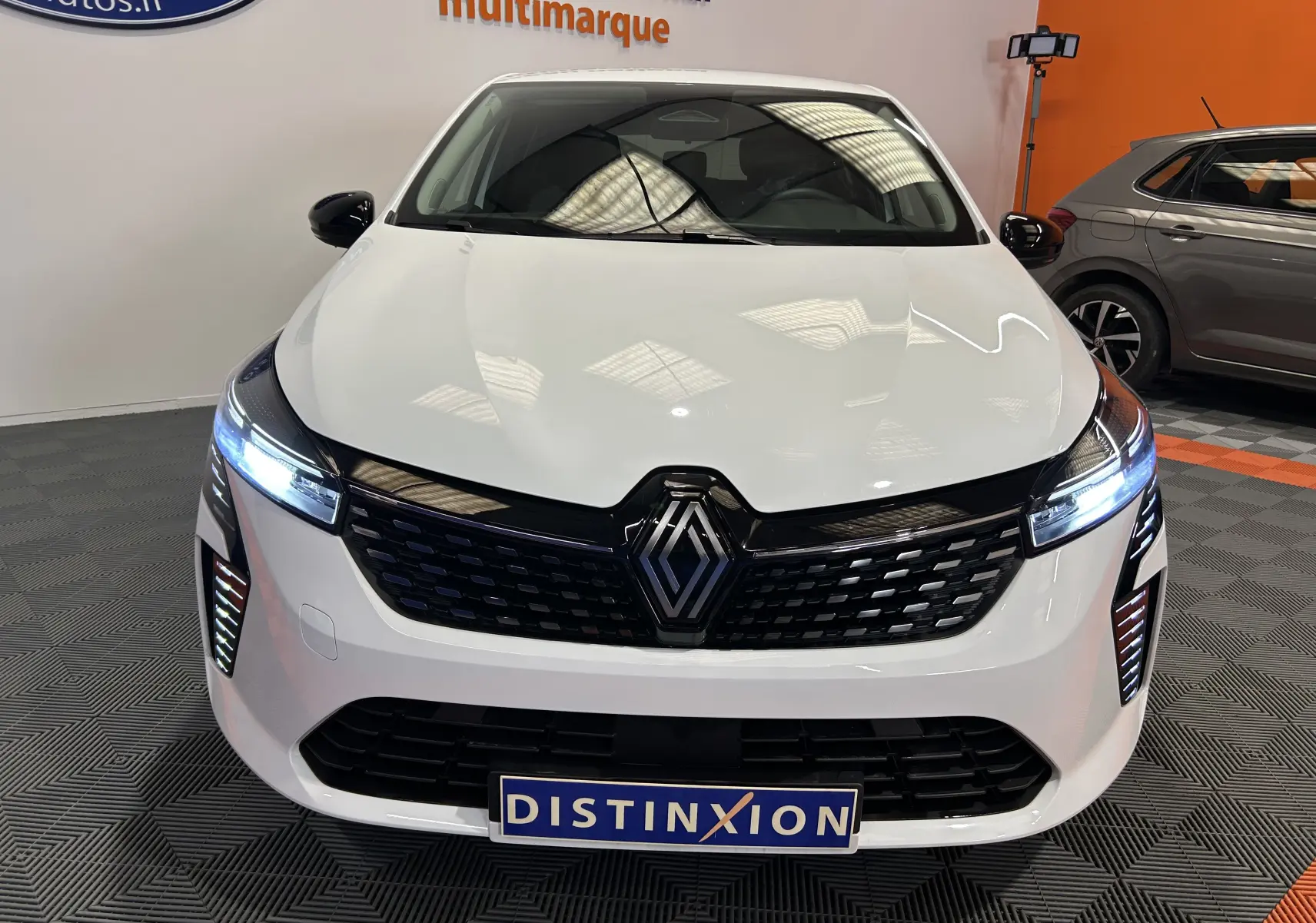 Vue avant d'une Renault Clio V blanche avec phares LED allumés et calandre noire dans un showroom.