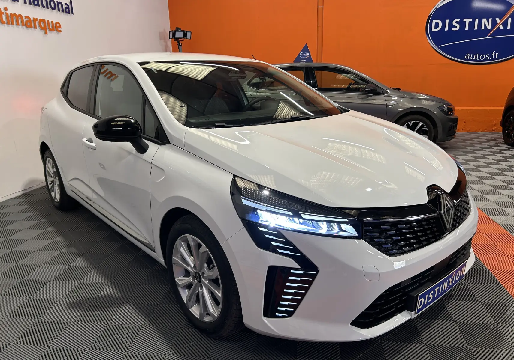 Renault Clio V blanche vue 3/4 avant droit, phares LED allumés et rétroviseurs noirs dans showroom.