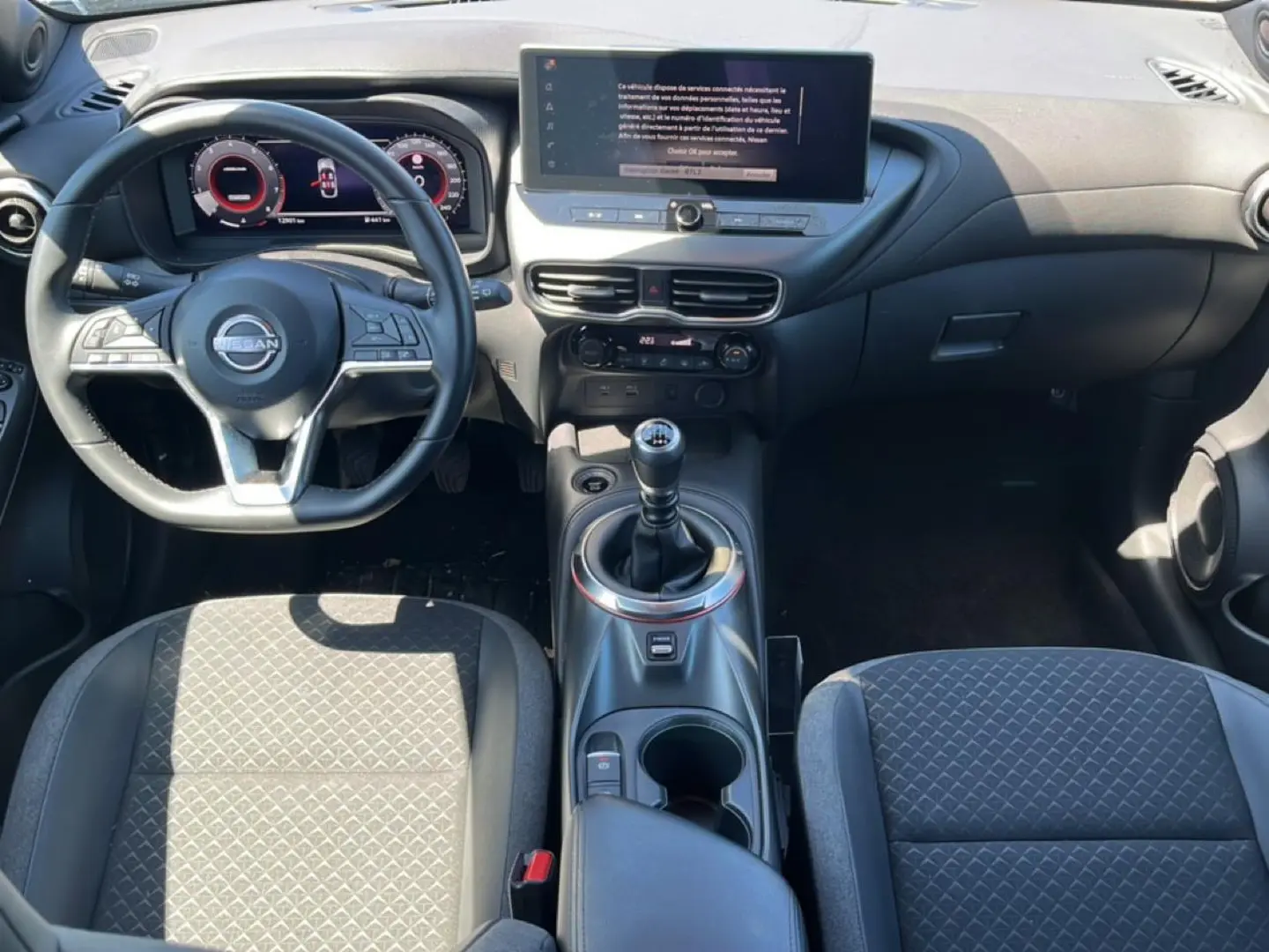 Vue intérieure avant du Nissan Juke 2025 gris, volant cuir multifonction et levier de vitesse manuel au centre.