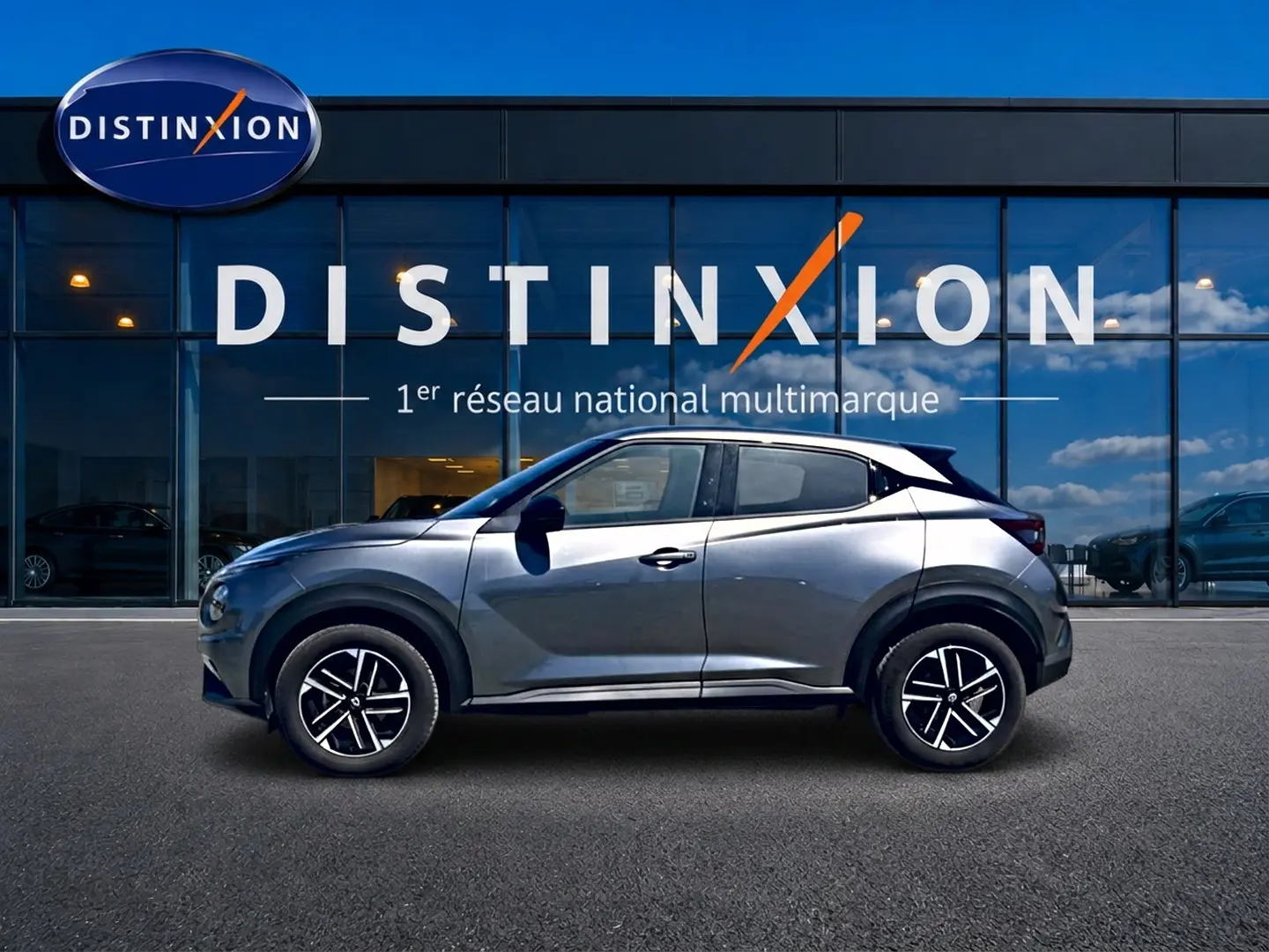 Profil droit d'un Nissan Juke gris 2025 garé devant une vitrine avec jantes alliage et toit noir contrasté.
