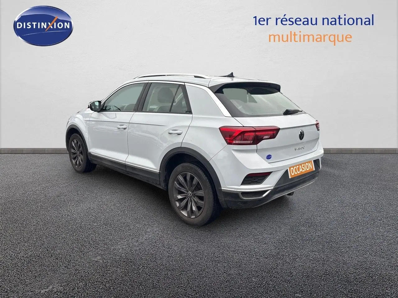 Volkswagen T-Roc blanc argenté vu en 3/4 arrière droit, avec jantes foncées et feux arrière allumés.