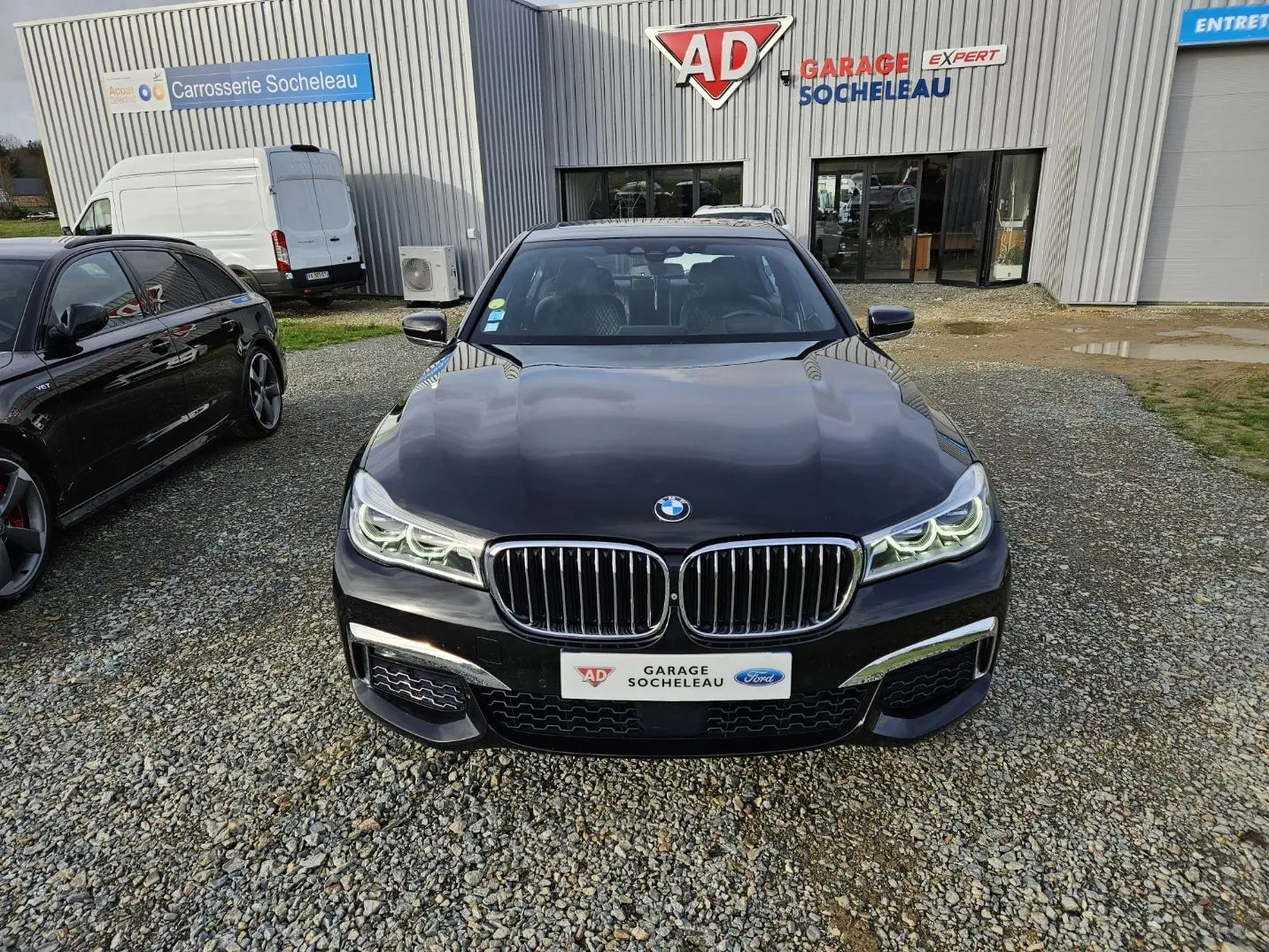BMW Série 7 G11 noire vue de face, avec calandre double haricot et phares LED allumés devant un garage.