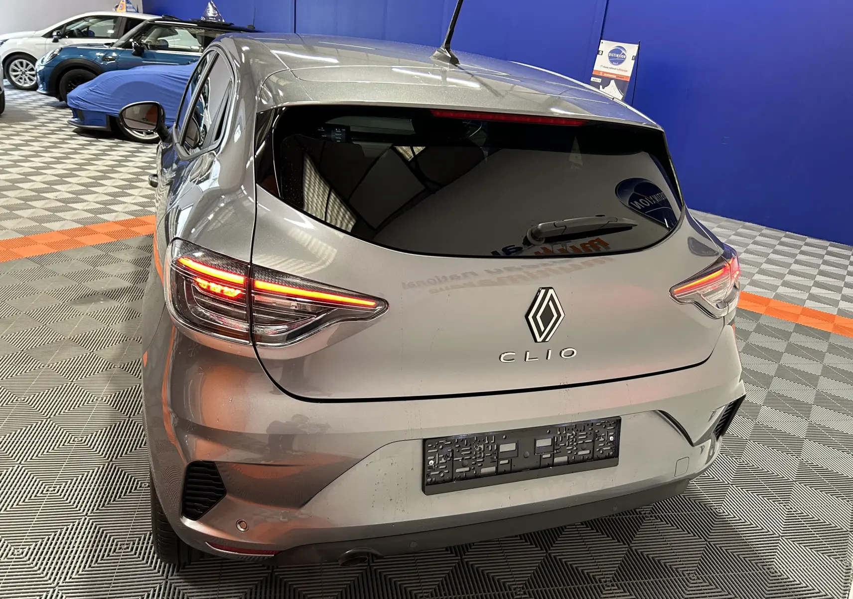 Vue 3/4 arrière droite d'une Renault Clio V gris schiste métal avec feux arrière allumés dans un showroom.