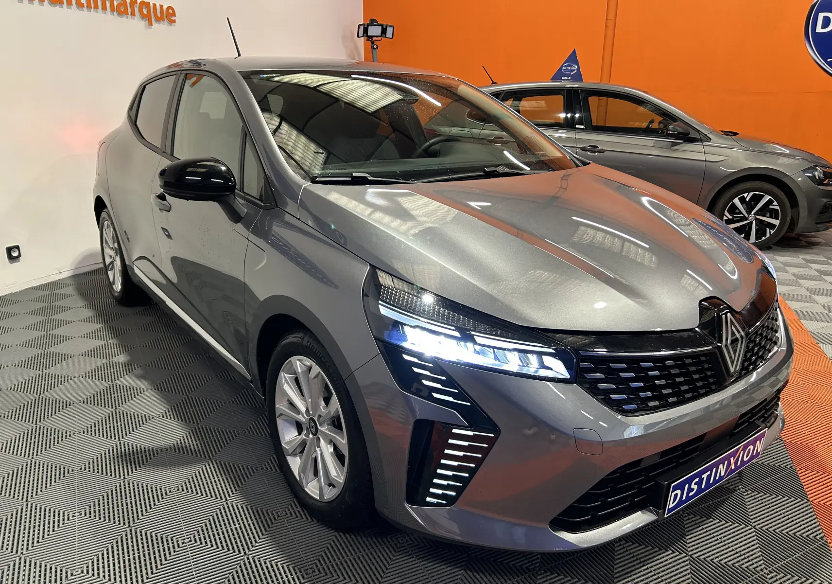 Renault Clio V gris schiste métal vue 3/4 avant droit avec phares LED allumés en intérieur showroom.
