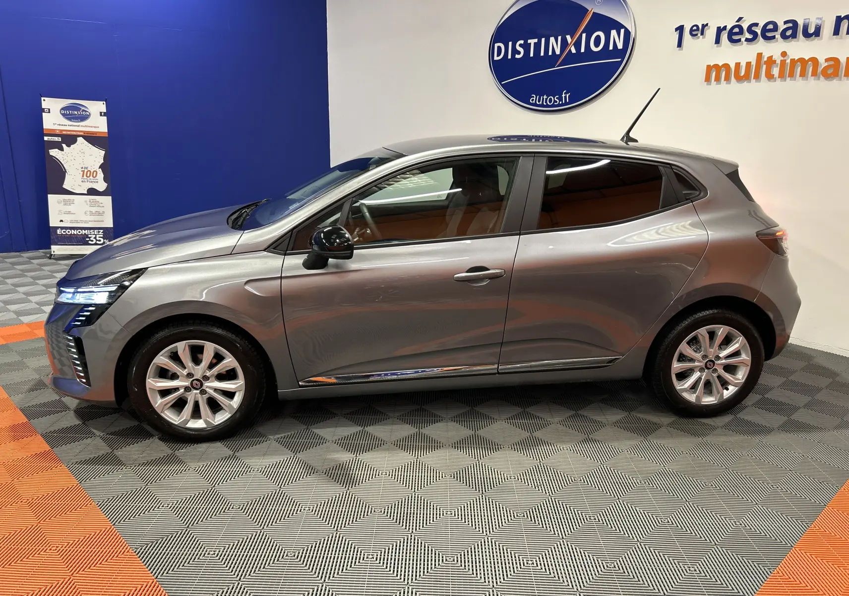 Profil côté gauche d'une Renault Clio V gris schiste métal, avec jantes alliage et feux avant LED allumés, en intérieur showroom.