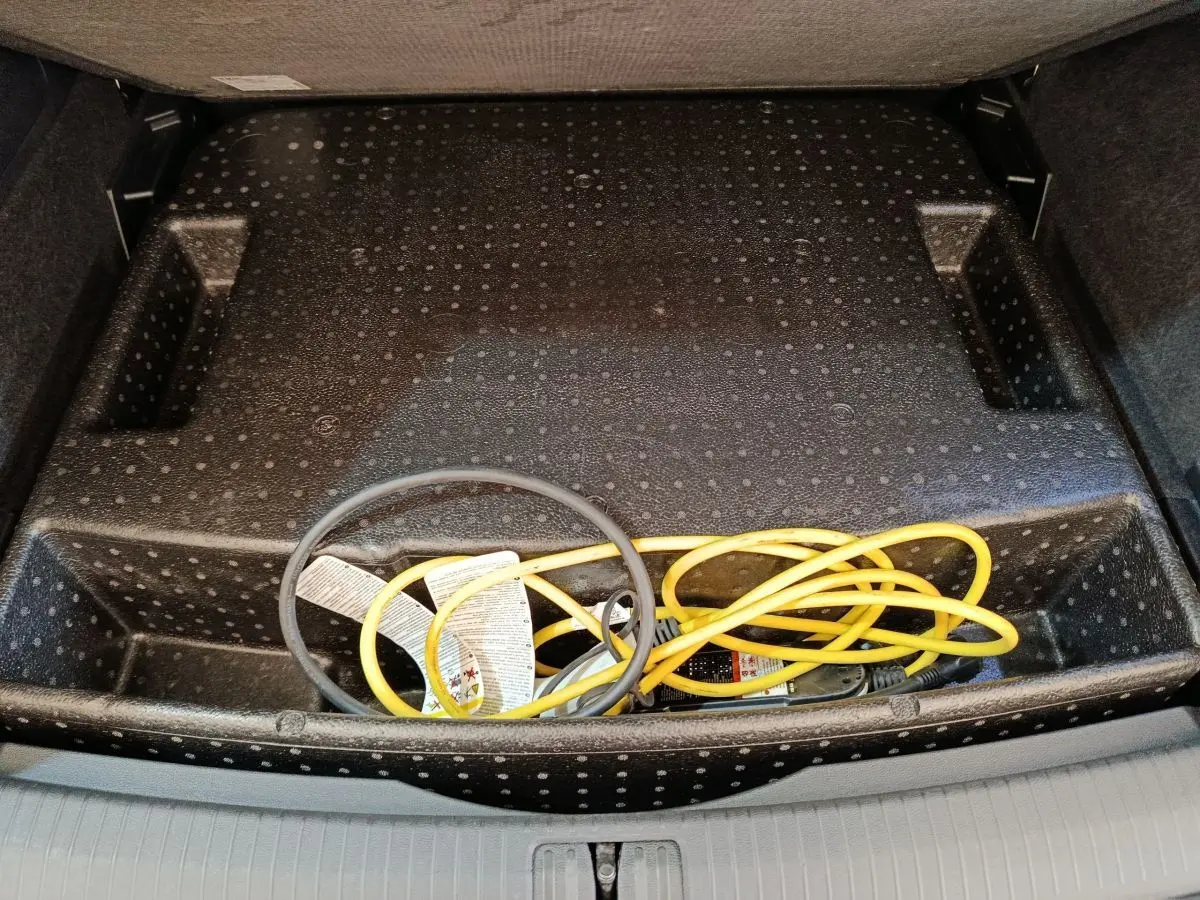 Vue plongeante sur le rangement sous plancher de coffre du Volkswagen Tiguan noir, avec câble de recharge jaune et noir visible.